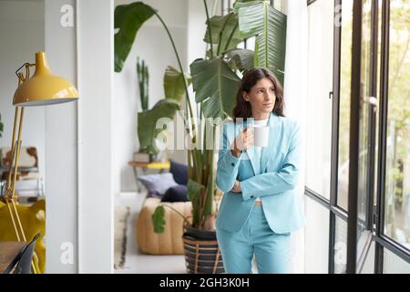 Geschäftsfrau, die Kaffee trinkt und sich aus dem Bürofenster lehnt. Eine inspirierende Frau schaut nachdenklich und reflektiv nach draußen, während sie eine Tasse hält. Stockfoto