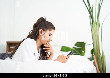Junge Latina-Frau, die ein Mobiltelefon auf dem Bett benutzt. Menschen, die von zu Hause aus verbunden sind, während sie sich entspannen. App, SMS, Online-Bildung, Telearbeit. Stockfoto