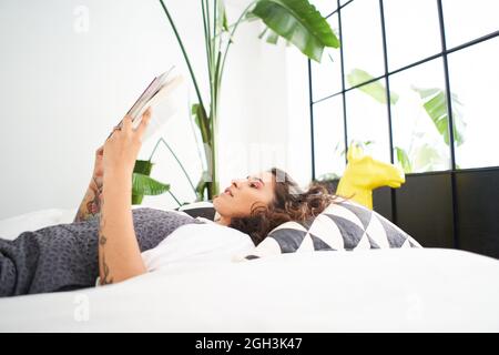 Junge Latina-Frau liest ein Buch, das auf dem Bett liegt. Menschen, die zu Hause studieren und lernen. Konzept von Freizeit und Kultur. Stockfoto
