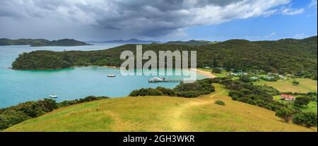 Panoramablick auf die Insel Urupukapuka in der Bay of Islands, Neuseeland. Ein Ausflugsboot wird am Kai festgebunden und eine Squall zieht vom Meer ein Stockfoto