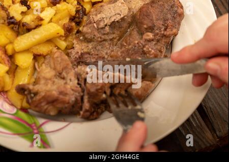 Jemand schneidet mit einem Messer und einer Gabel ein Stück saftiges Braten auf einem Teller gebratener Kartoffeln. Nahaufnahme Selektiver Fokus Stockfoto