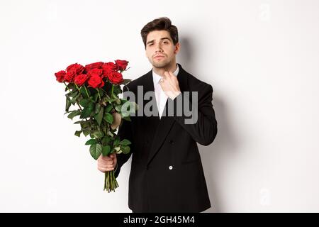 Bild von eleganten und frechen Mann in schwarzem Anzug, selbstbewusst und hält Strauß von roten Rosen, gehen auf ein romantisches Date, stehen gegen weiß Stockfoto