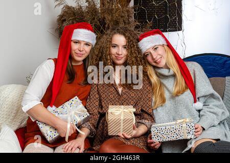 Drei glückliche junge Frauen in roten Zwerghüten sitzen auf der Couch und halten Weihnachtsgeschenke Stockfoto