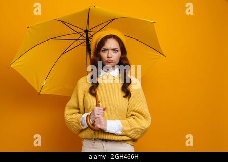 Glückliche junge Frau mit langen roten Haaren trägt Baskenmütze und Pullover unter Regenschirm auf gelbem Hintergrund Stockfoto