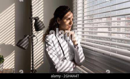 Lächelnde, nachdenkliche Ärztin, die im Büro am Fenster steht Stockfoto
