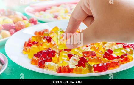 Hand nimmt bunte Gummibärchen Bonbons des Tellers. Nahaufnahme. Gelee-Leckereien für Kinder und Erwachsene. Stockfoto