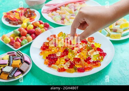 Gummis, Gelee, Lakritz-Bonbons auf dem Tisch. Die Hand nimmt einen bunten Gummibären von einem Teller. Eine Auswahl an Leckereien für den Urlaub. Stockfoto