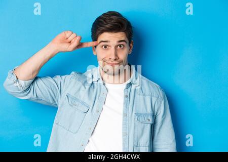 Nahaufnahme eines Mannes, der sich dumm oder verrückt verhält, mit dem Finger auf dem Kopf rollt und auf die Kamera schaut, die auf blauem Hintergrund steht. Stockfoto