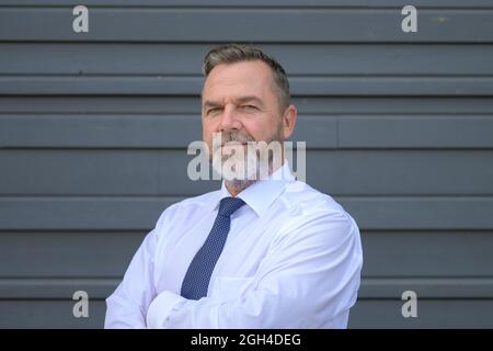 Ein Geschäftsmann, der die Kamera mit einem ernsthaften, einschätzen Blick untersucht, während er mit gefalteten Armen vor einer grauen Außenwand steht Stockfoto