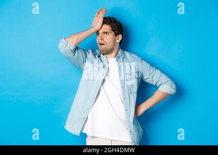 Verärgert Mann macht Facepalm und schaut besorgt weg, vergessen etwas wichtiges, schlagen Stirn beunruhigt, stehen vor blauem Hintergrund. Stockfoto