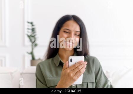 Schöne junge indische Frau mit Smartphone sitzt auf dem Sofa zu Hause. Die charmante, lächelnde Mixed-Race-Frau hält das Telefon und schaut auf den Bildschirm, macht Videoanruf, schaut sich Videos an, verbringt Freizeit im Internet Stockfoto