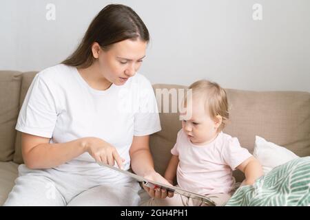 Mutter erklärt Dinge, während blondes Mädchen Buch ansieht. Sitzen auf einem Sofa im Wohnzimmer Stockfoto