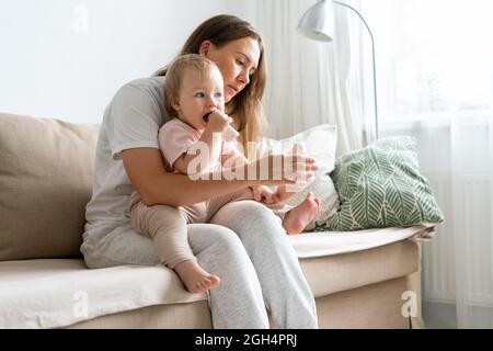 Lächelndes, hellhaariges Kindermädchen, das zu Hause auf der blonden blauäugigen Mädchencouch sitzt und Socken setzt Stockfoto