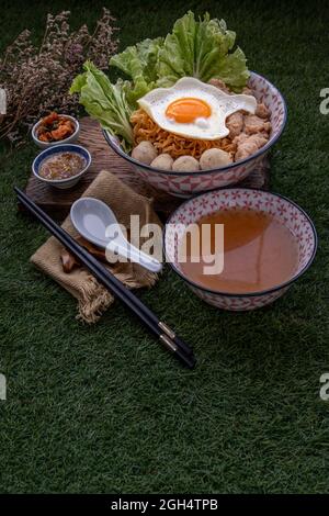 Eiernudeln mit Schweinefleischkugel, Hackfleisch, Spiegelei, Gemüse und Suppen in Schüssel und Essstäbchen mit Löffel und Holz Hintergrund kochen. Thailändische Nudelsuppe Stockfoto