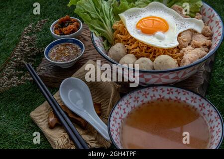 Eiernudeln mit Schweinefleischkugel, Hackfleisch, Spiegelei, Gemüse und Suppen in Schüssel und Essstäbchen mit Löffel und Holz Hintergrund kochen. Thailändische Nudelsuppe Stockfoto