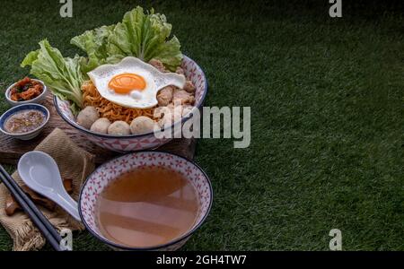 Eiernudeln mit Schweinefleischkugel, Hackfleisch, Spiegelei, Gemüse und Suppen in Schüssel und Essstäbchen mit Löffel und Holz Hintergrund kochen. Thailändische Nudelsuppe Stockfoto