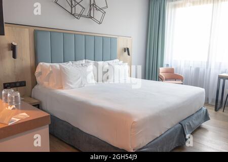 Helles und gemütliches, modernes Hotelzimmer mit komfortablem Kingsize-Bett, großem Fenster und blauem Kopfteil. Stockfoto