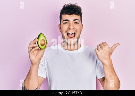 Junger hispanischer Mann, der die mittlere Avocado hält und den Daumen zur Seite zeigt, lächelt glücklich mit offenem Mund Stockfoto