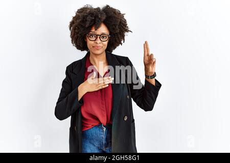 afroamerikanische Frau mit Afro-Haaren, die eine Business-Jacke und eine Brille trägt, die lächelnd mit der Hand auf der Brust und den Fingern nach oben flucht, was ein Loyalitätsversprechen macht Stockfoto