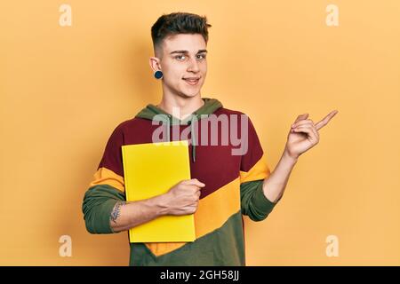 Junge kaukasische Junge mit Ohren Erweiterung halten Buch lächelnd glücklich zeigt mit Hand und Finger zur Seite Stockfoto