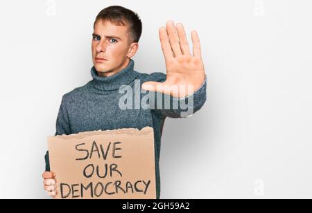 Junger blonder Mann hält unser Demokratie-Protestbanner mit offener Hand und tut Stoppschild mit ernstem und selbstbewusster Ausdruck, Verteidigungsgeste Stockfoto