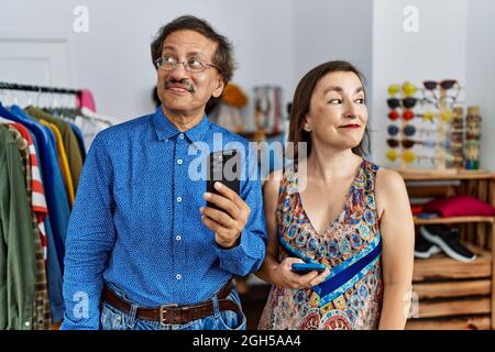 Interrassisches Paar im mittleren Alter im Einzelhandelsgeschäft mit einem Smartphone, das lächelt und zur Seite schaut und denkend wegstarrt. Stockfoto