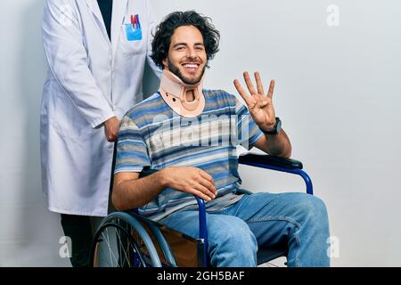 Gutaussehender hispanischer Mann, der auf dem Rollstuhl sitzt und einen Halsband trägt und mit den vier Fingern nach oben zeigt, während er selbstbewusst und glücklich lächelt. Stockfoto