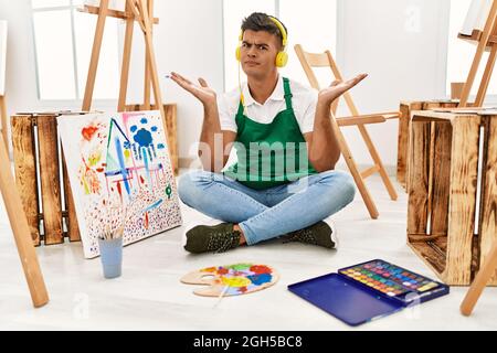 Junger hispanischer Mann im Kunststudio ahnungslos und verwirrt Ausdruck mit Armen und Händen angehoben. Zweifel Konzept. Stockfoto