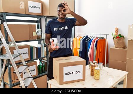 Junger afroamerikanischer Freiwilliger, der Spendenbox für wohltätige Zwecke verpackt und dabei mit lächelnder Hand und einem glücklichen Gesicht durch die Finger schaut, eine gute Geste tut Stockfoto