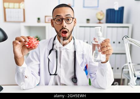 afroamerikanischer Arzt, der ein Virus-Spielzeug hält, ein Desinfektionsmittel-Gel im Schockgesicht, skeptisch und sarkastisch aussehend, überrascht mit offenem Mund Stockfoto
