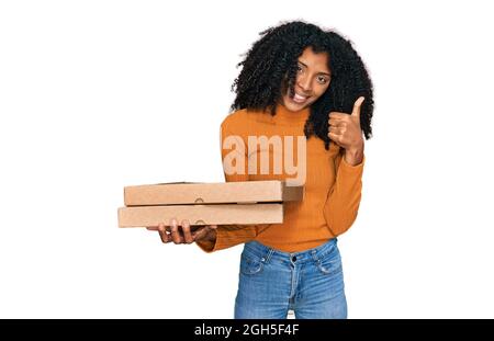 Junge afroamerikanische Mädchen hält Lieferung Pizza Box lächelnd glücklich und positiv, Daumen nach oben tun ausgezeichnet und Zustimmung Zeichen Stockfoto