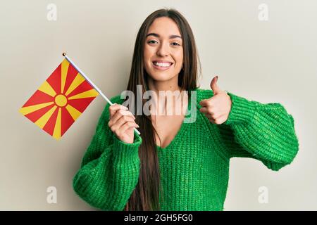 Junge hispanische Mädchen hält mazedonische Flagge lächelnd glücklich und positiv, Daumen nach oben tun ausgezeichnet und Zustimmung Zeichen Stockfoto