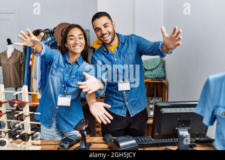 Junge interrassische Menschen, die in einer Einzelhandelsboutique arbeiten, blicken auf die Kamera und lächeln mit offenen Armen zur Umarmung. Fröhlicher Ausdruck, der Glück umarmt. Stockfoto