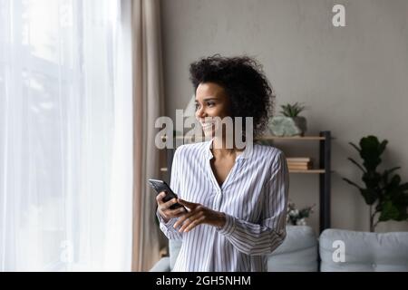 Fröhliche junge schwarze Frau mit Telefon in der Nähe des Fensters Stockfoto