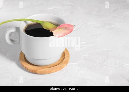 Frischer Kaffee in einer weißen Tasse mit einer zartrosa Calla-Blume auf hellgrauem Tischhintergrund, romantischer Morgenhintergrund Stockfoto