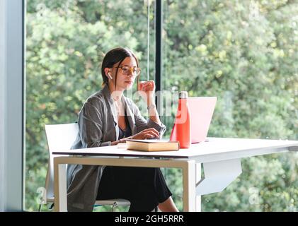 Seitenansicht einer Studentin, die auf Netbook tippt, während sie mit dem Buch am Tisch sitzt und Hausaufgaben in der Universitätsbibliothek macht Stockfoto
