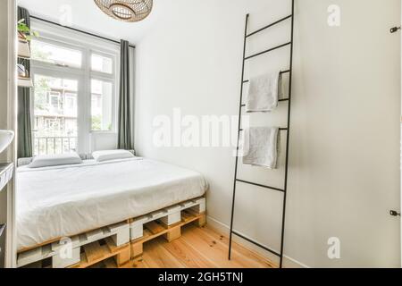 Leiter mit Handtüchern in der Nähe des Palettenbetts in einem hellen, minimalistischen Schlafzimmer mit grauen Wänden am Tag Stockfoto