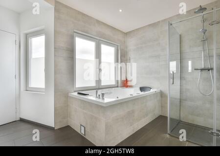 Stilvolles Badezimmer-Interieur mit grauen Marmorfliesen und Fenstern ausgestattet mit Dusche und luxuriöser Badewanne in modernem Apartment Stockfoto