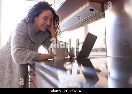 Geschäftsfrau mit lockigen Haaren in der Küche, die eine Infusion nahm, während sie ihren Laptop benutzte und zu Hause arbeitete Stockfoto