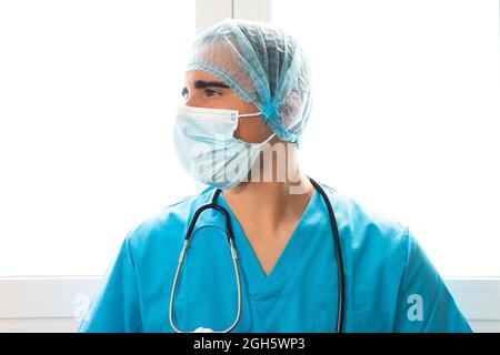 Seitenansicht des männlichen Sanitäter in Maske und Uniform, die in der Klinik in der Nähe des Fensters steht Stockfoto