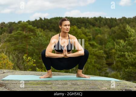 Barfuß lange junge Frau, die Malasana auf der Matte posiert und wegschaut, während sie im Sommer Yoga auf dem Fels in der Natur praktiziert Stockfoto