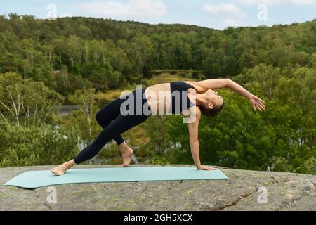 Schlanke, anmutige Frau in schwarzen Leggings und BH, die in Wild Thing Pose balancieren und den Körper während der Yoga-Sitzung gegen grüne Bäume auf dem Land strecken Stockfoto
