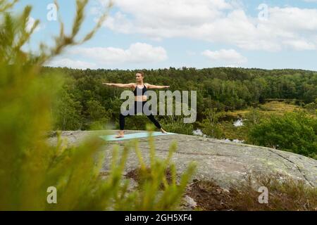 Barfuß-Mädchen in voller Länge, die Warrior auf der Matte posiert und wegschaut, während sie im Sommer Yoga auf dem Fels in der Natur praktiziert Stockfoto