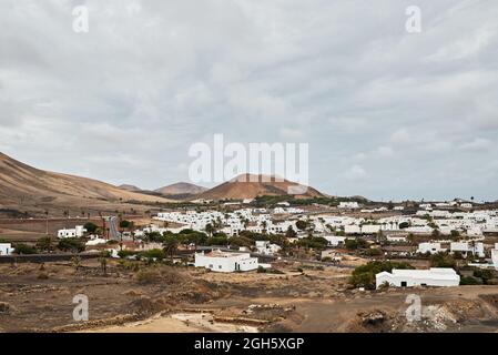 Weiße Häuser und grüne Bäume auf der Straße der Stadt in der Nähe von trockenen Hügeln gegen grau bewölkten Himmel in Fuerteventura, Spanien Stockfoto