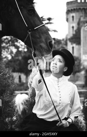 Serious adult schwarz weiblich in Hut und elegantes Outfit mit Handschuhen streicheln braunes Pferd in der Nähe von grünen Pflanzen und Bäumen auf dem Land Stockfoto