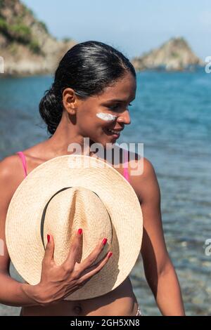 Verträumte, ethnische Touristin mit Sonnencreme auf den Wangen und Nase, die mit einem Hut in der Hand im Sonnenlicht gegen das Meer steht Stockfoto