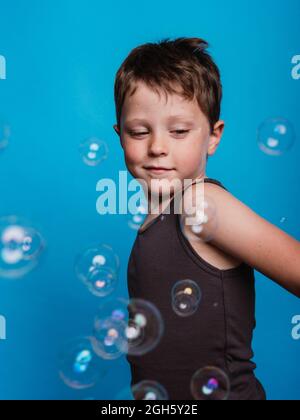 Vorteen Junge schaut weg im Studio mit fliegenden Seifenblasen auf blauem Hintergrund Stockfoto