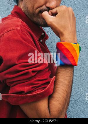 Zugeschnittenes, nicht erkennbares, heiteres, ethnisch homosexuelles Männchen mit Regenbogenbandana auf der Hand, das Bizeps auf dem Hintergrund einer grauen Mauer in der Stadt zeigt Stockfoto