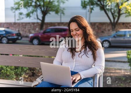 Fröhliche junge lateinamerikanische Studentin, die am Sommertag auf einer Holzbank auf der Straße der Stadt auf einem Laptop surft Stockfoto