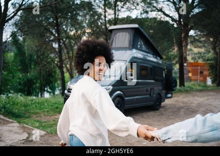 Lächelnde afroamerikanische Frau, die die Hände eines beschnittenen Freundes hält, die Hände hält, während sie spazieren geht und gegen den Camper schaut Stockfoto
